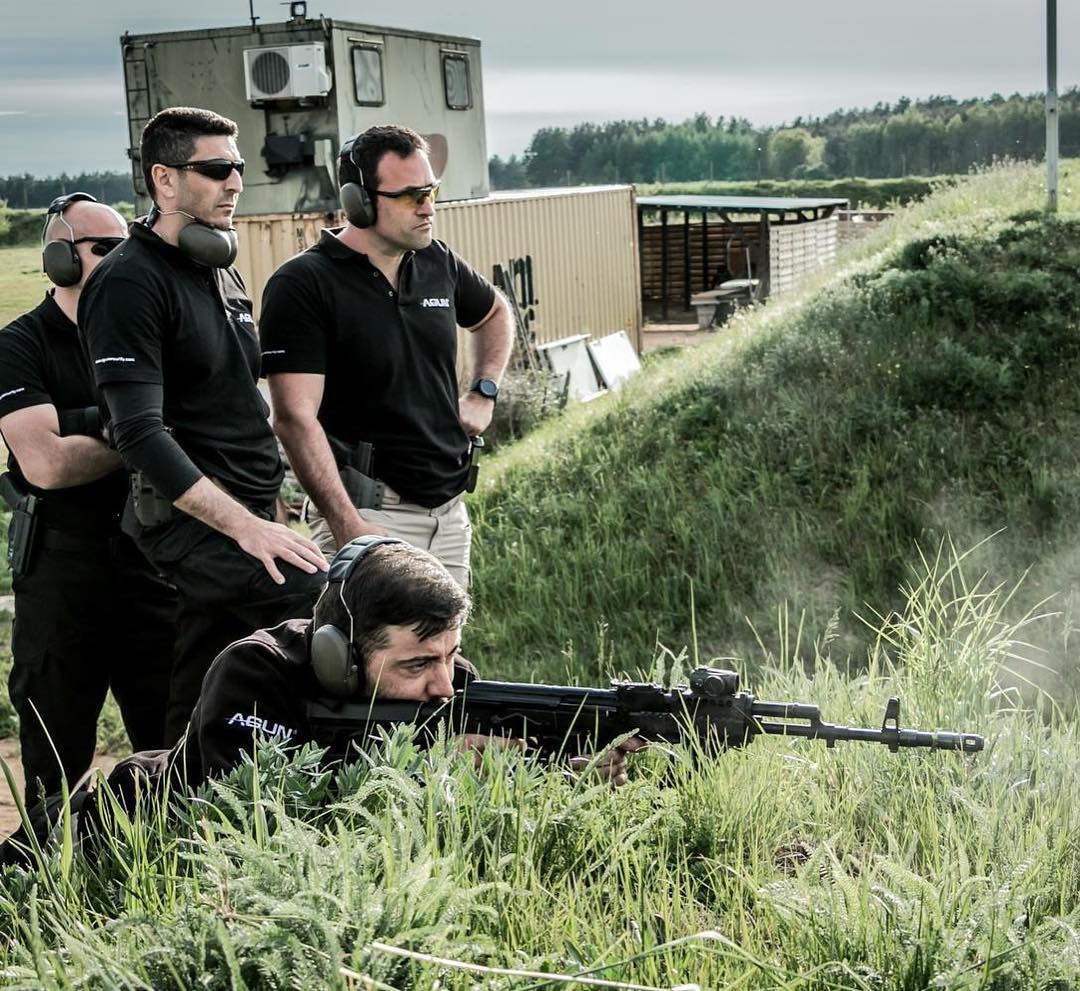 Instructors standing over a trainee firing a rifle from the prone position in long grass