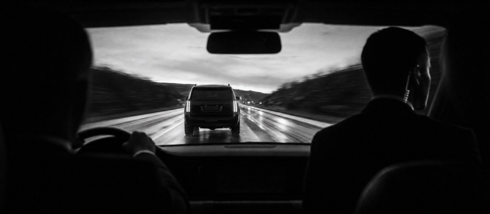 Security convoy on a wet road at dusk, an armoured SUV seen from the follow car